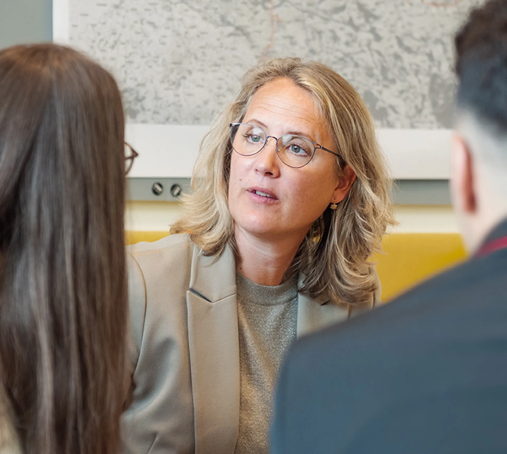 Drei Personen sitzen beisammen und unterhalten sich. Die mittlere Person hat schulterlanges blondes Haar und trägt einen grauen Blazer über einem grauen Oberteil. Die Gesichter der anderen Personen sind nicht zu sehen, da sie von hinten fotografiert wurden. Es handelt sich links um Person mit langen, brauen Haaren und einer Brille und rechts um eine Person mit kurzen, dunklen Haaren, die ein Sakko trägt. Im Hintergrund hängt eine großformatige Karte an der Wand.
