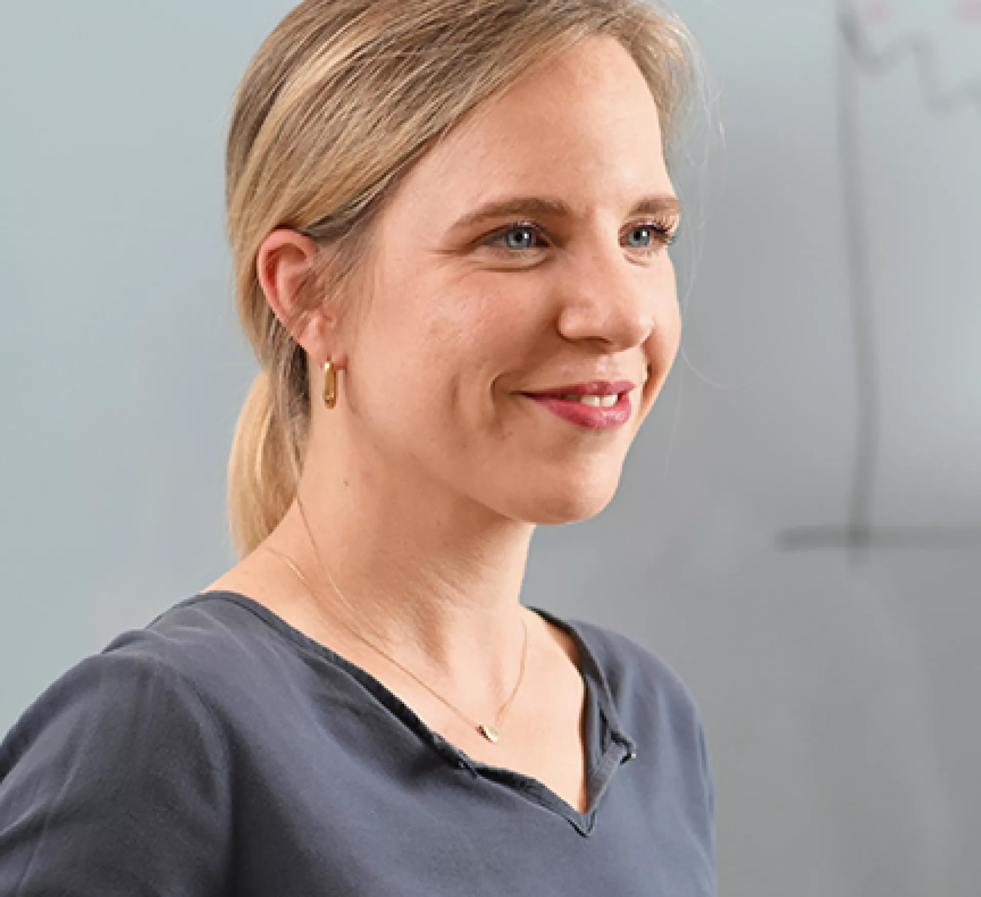 Eine lächelnde Person mit einem mittellangem, zu einem glatten Pferdeschwanz gebundenem, blonden Haar trägt ein dunkles Oberteil und eine Halskette. Im Hintergrund ist eine weiße Tafel mit einer gezeichneten Grafik zu sehen.