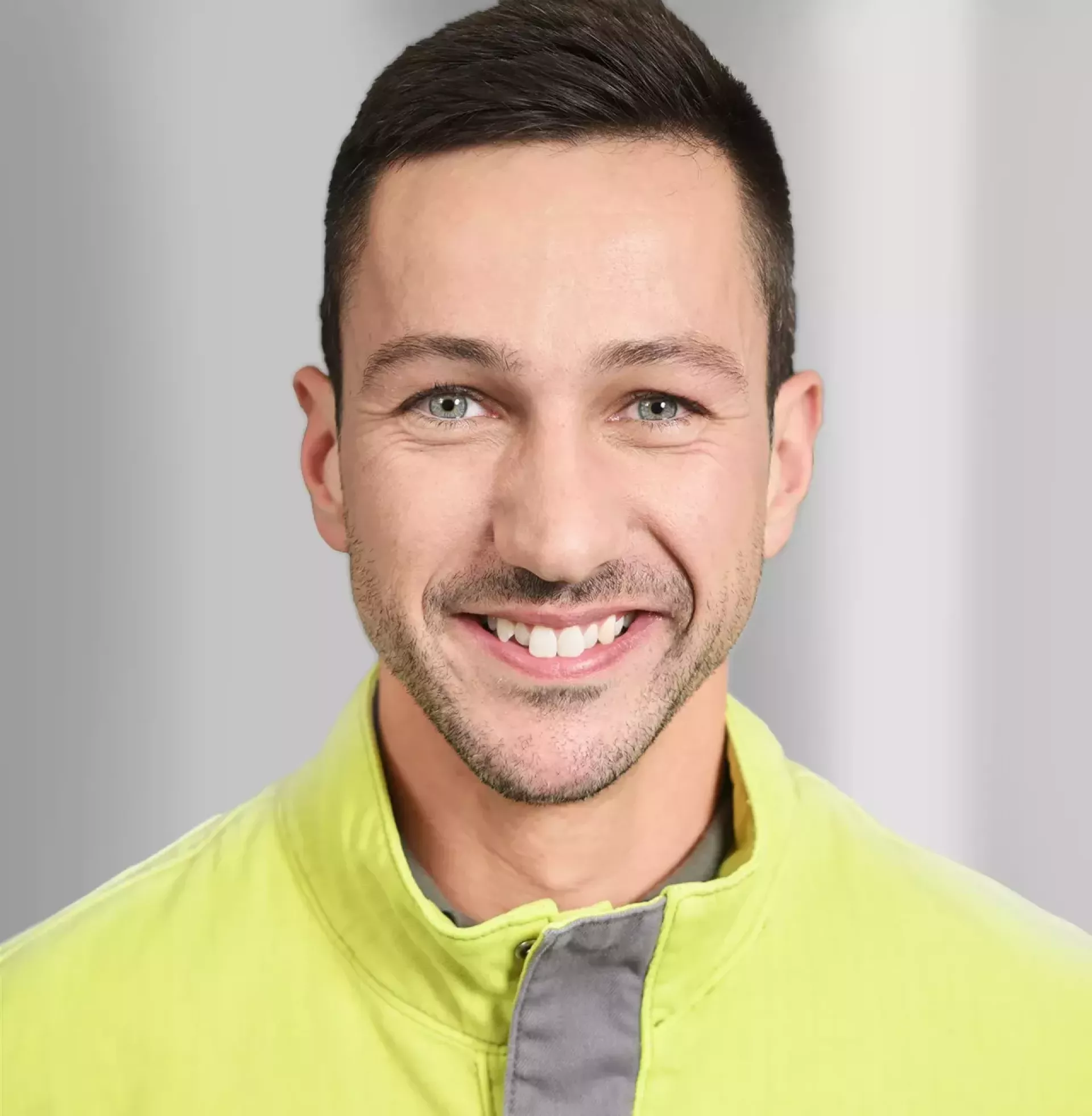    Porträt einer Person mit kurzen, dunkelblonden Haaren und Brille, die eine gelbe Warnweste mit hohem Kragen trägt. Der Hintergrund ist einfarbig grau und schlicht, ohne erkennbare Objekte oder Strukturen. 