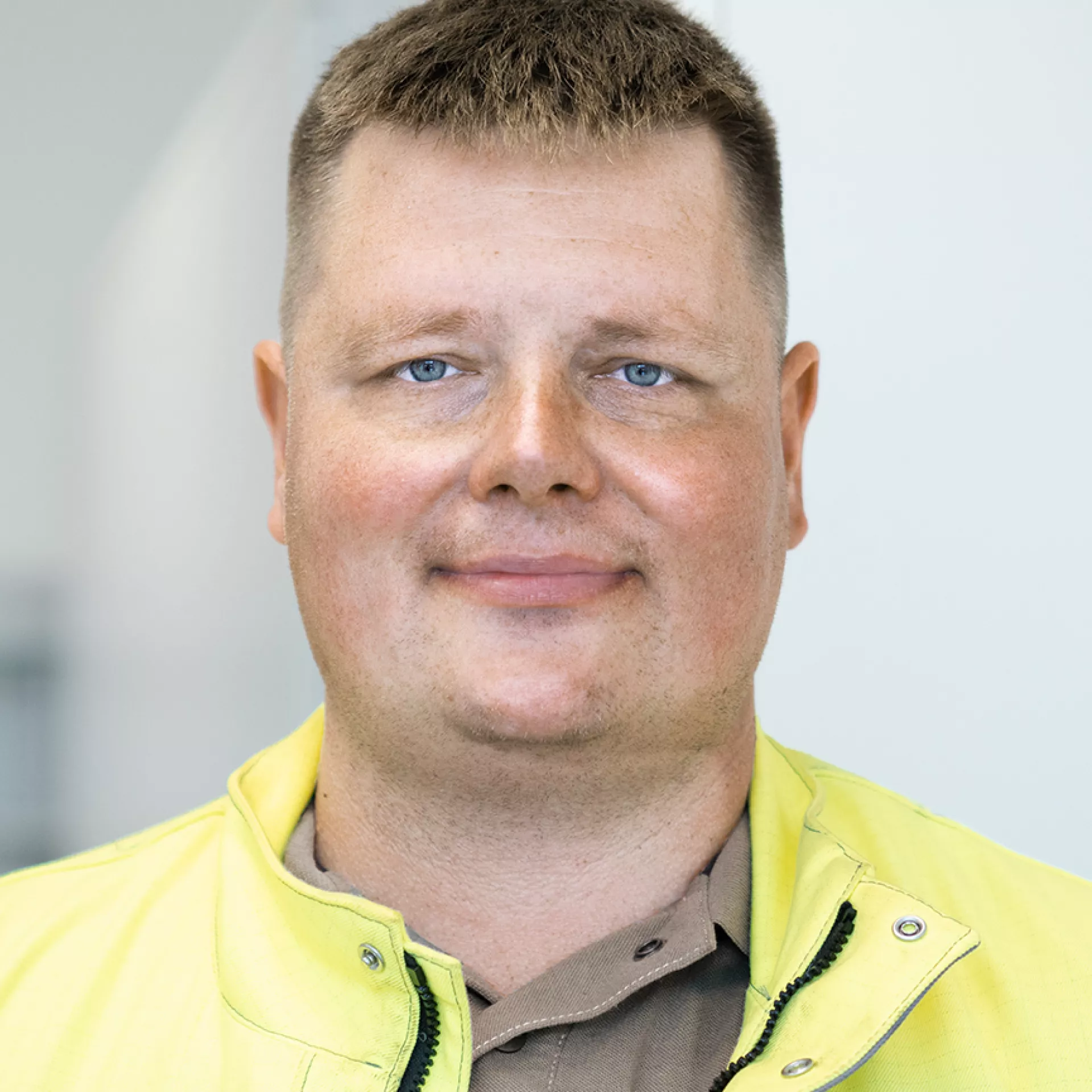 Eine Person mit kurzen, braunen Haaren trägt eine persönliche Schutzausrüstung (gelbe Jacke mit Reißverschluss und Druckknöpfen sowie ein braunes Hemd darunter). Der Hintergrund ist unscharf und zeigt eine helle Umgebung.