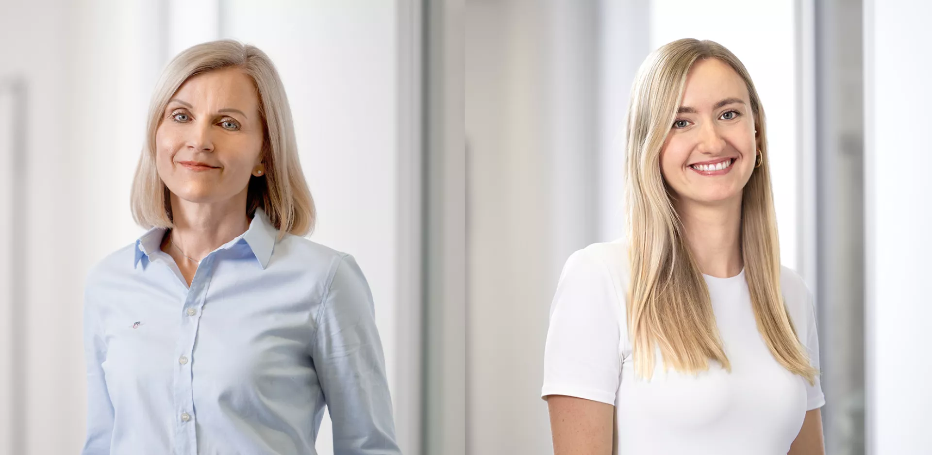 Zwei Personen befinden sich in einem modernen, hellen Bürogebäude. Die linke Person trägt eine hellblaue, langärmelige Bluse und hat schulterlanges, blondes Haar. Die rechte Person trägt ein weißes, kurzärmeliges T-Shirt und hat ebenfalls langes, blondes Haar, das über die Schultern fällt. Der Hintergrund ist unscharf und es ist eine helle Büroumgebung zu sehen. 